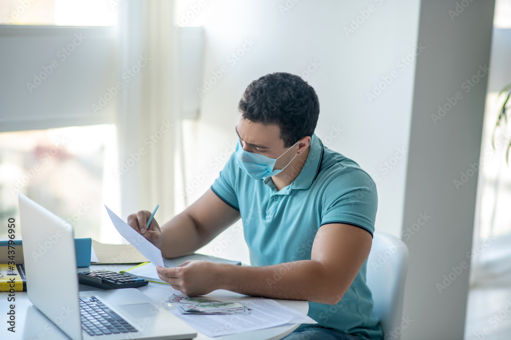 Sad dark-haired male sitting at his desk in protective mask, writing