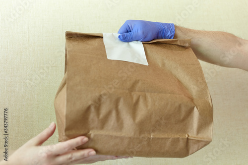 Male hands in blue latex glove gives paper bag to female hands. Closeup of male hands in gloves hold brown craft paper bag with blank white sticker. Delivery service concept. Copy space area.