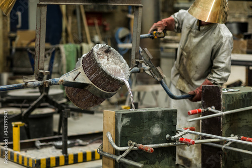 A craftsmen in silver protective clothing performing a metal sand casting technique by pouring molten aluminum silver colored liquid into a mold in a industrial environment, workshop space photo