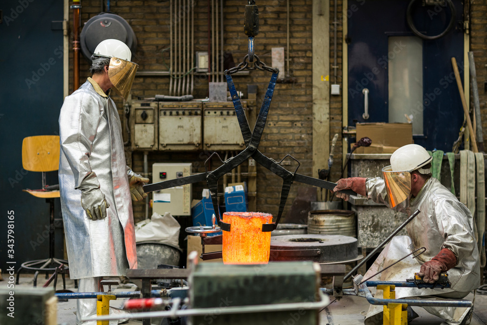 Two craftsmen in silver protective clothing doing metal sand casting technique pouring molten aluminum silver colored liquid into a mold in a industrial environment, workshop space Two craftsmen in silver protective clothing doing metal sand casting technique pouring molten aluminum silver colored liquid into a mold in a industrial environment, workshop space