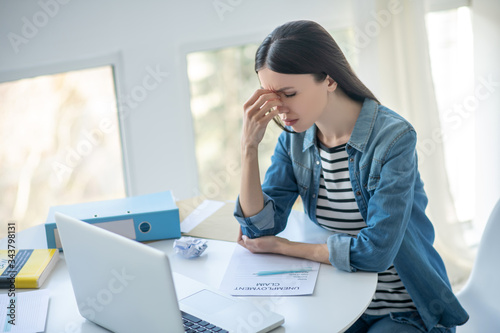 Upset dark-haired female sitting at her desk with unemployment claim, touching her nose bridge photo