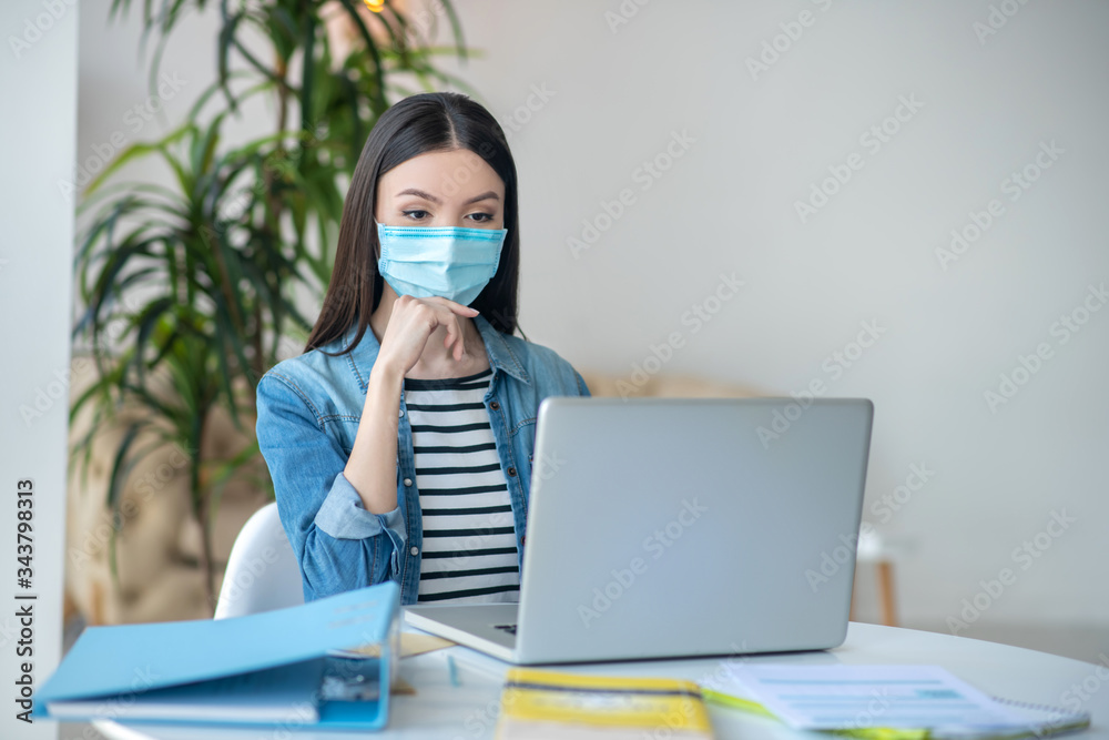 Dark-haired female sitting at her desk. looking at laptop, wearing protective mask Dark-haired female sitting at her desk. looking at laptop, wearing protective mask