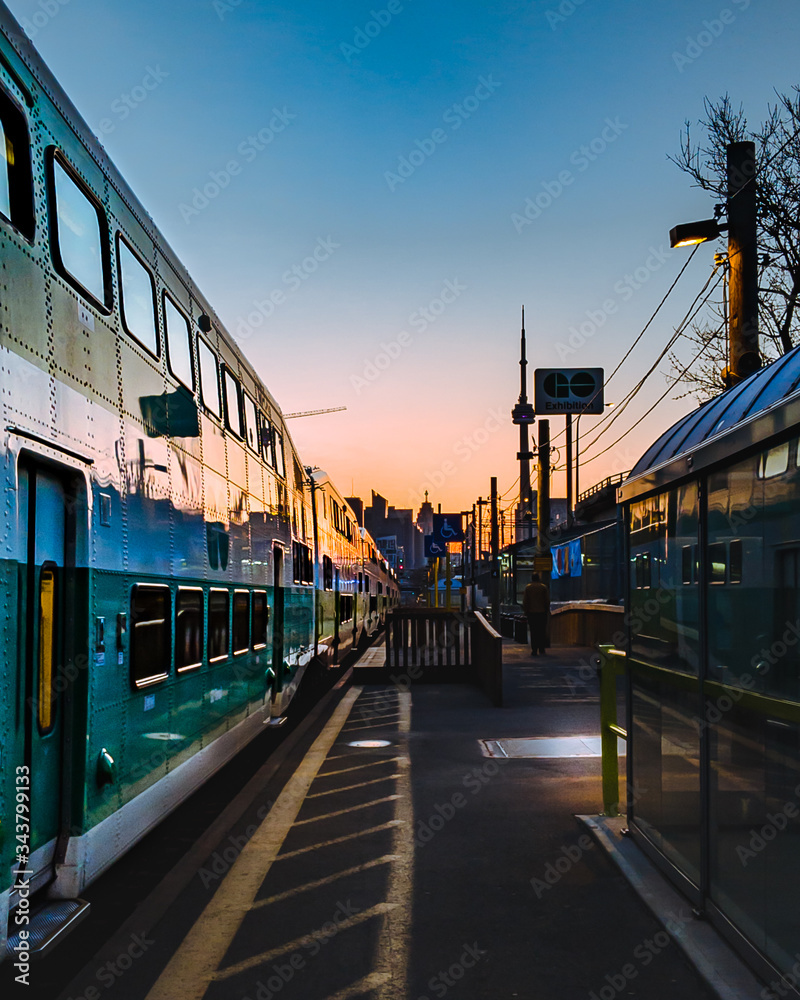 A green and white commuter train waits for passengers at a rail station platform early in the morning with the rising sun in the background A green and white commuter train waits for passengers at a rail station platform early in the morning with the rising sun in the background
