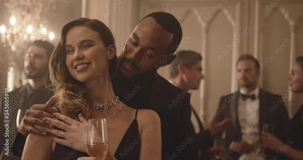 Handsome young man dancing with a beautiful woman at party with people in background discussing. Loving couple dancing at gala night.

