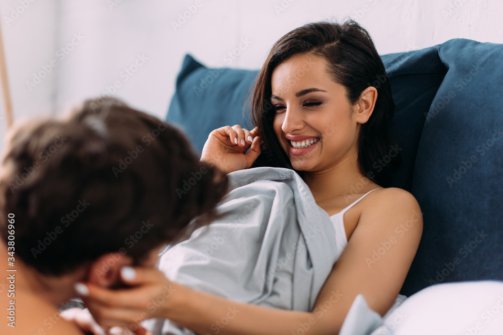 Selective focus of happy woman smiling, touching boyfriend and lying under blanket on bed in bedroom Selective focus of happy woman smiling, touching boyfriend and lying under blanket on bed in bedroom