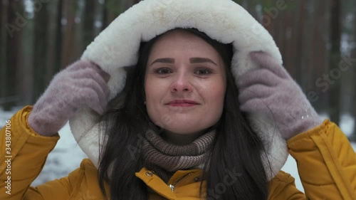 Winter Portrait of a young woman in a yellow jacket standing in a snow-covered forest and looking at the camera. A woman in a snowfall puts on the hood of her jacket and smiles.