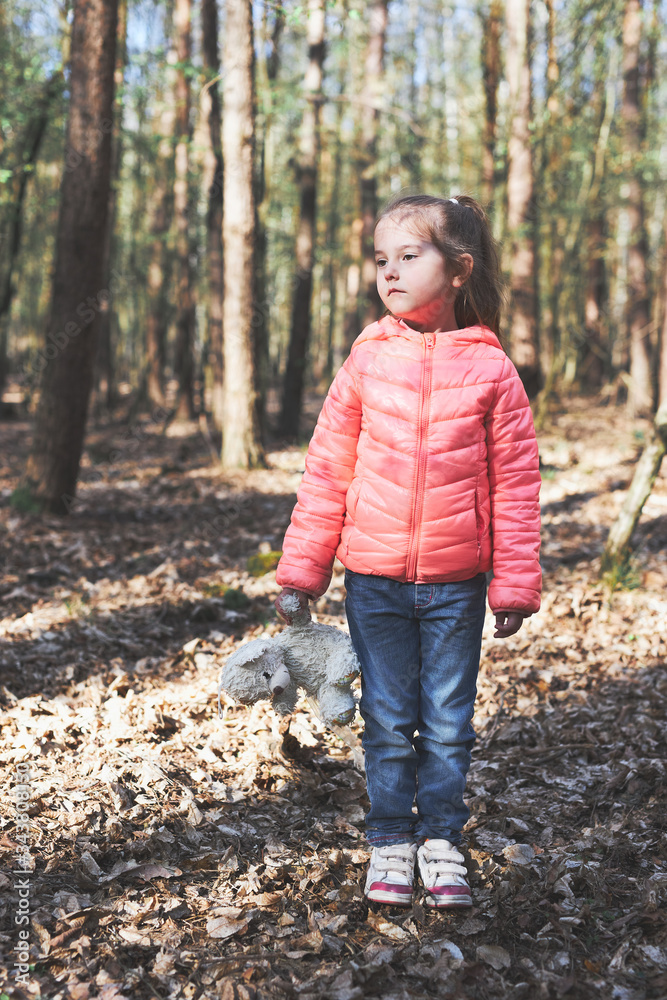 Little girl child standing on stump in a forest during a walk on sunny spring day keeping a toy teddy bear looking away Little girl child standing on stump in a forest during a walk on sunny spring day keeping a toy teddy bear looking away