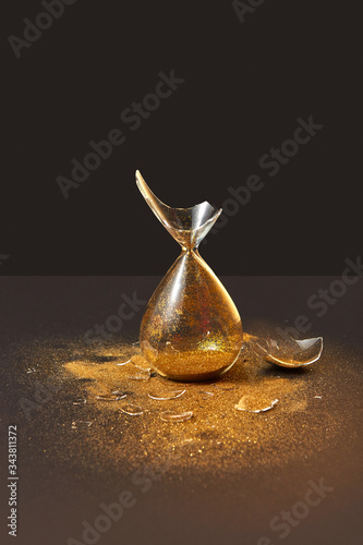 Fotografie Cracked antique hourglass with golden sand on a black duotone background