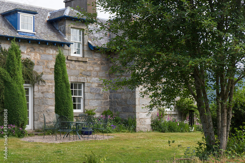 Garden of an English manor house in gray stone, with large white wooden windows, two cypresses on the sides of the door, baked iron garden furniture