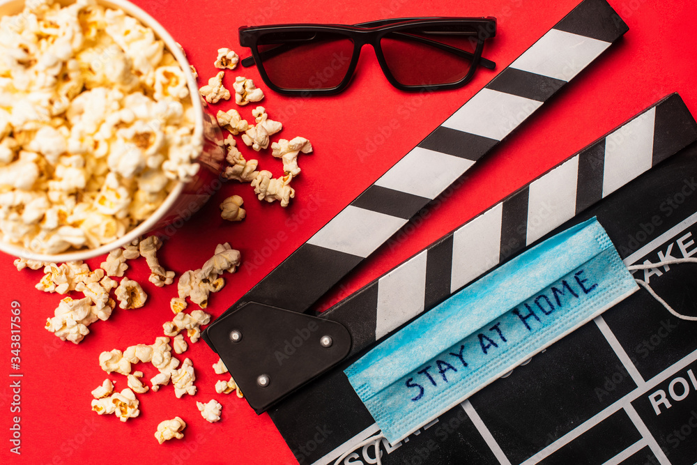 Top view of clapperboard with medical mask and stay at home lettering near popcorn and sunglasses on red surface