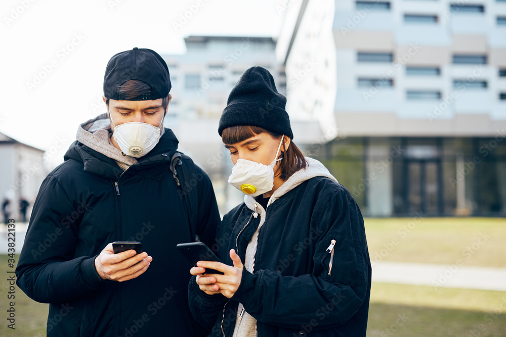 young couple in casual clothes wearing protection masks and using their phones on the street