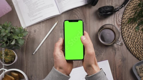 Phone with green screen chroma key in male hands. Thumb swipe left and. Top view. Workplace on wooden table with many things on the background. Daylight. POV. 4k video.