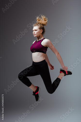 curly and sportive woman in sportswear jumping on grey