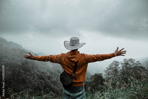 Hombre de espaldas con sombrero blanco y una sudadera naranja, abriendo los brazos a la naturaleza 