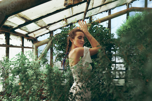 A young beautiful brunette woman in white dress standing in a greenhouse between green plants with lush foliage; dancing in orangery