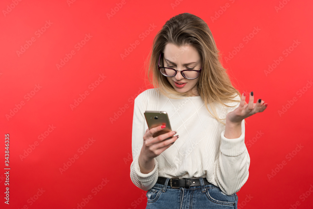 Young girl with glasses with blond hair looks into the smartphone with misunderstanding, she gesticulates her hand, stands on a red background Young girl with glasses with blond hair looks into the smartphone with misunderstanding, she gesticulates her hand, stands on a red background