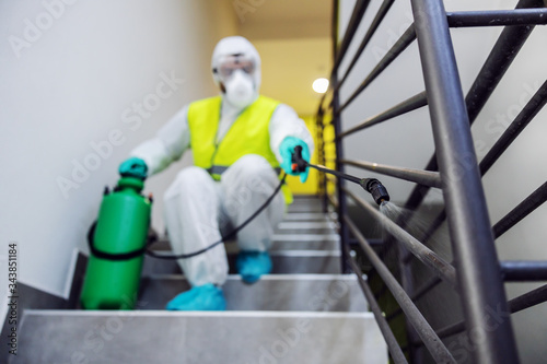 Closeup of worker in sterile suit and vest sterilizing railing of a building from corona virus / covid 19. Selective focus on sprayer.