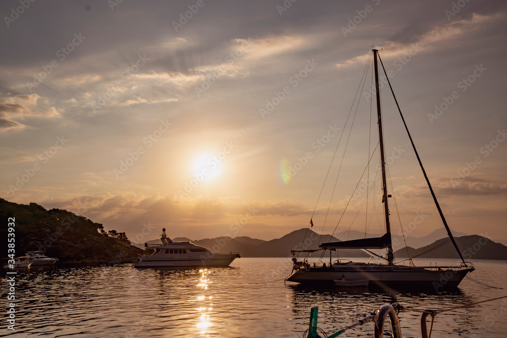 A bay at the Aegean sea with anchoring luxury sailing yachts at sunset. Misty sky with clouds and sun reflecting in water with copy space
