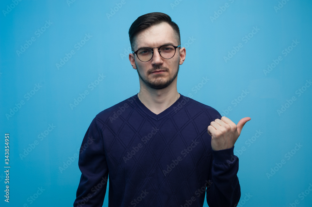 Positive young man in casual clothes and glasses points with his finger something useful on blue background Positive young man in casual clothes and glasses points with his finger something useful on blue background