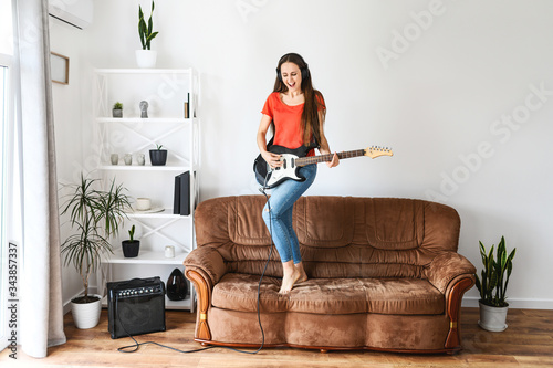 Fun at home on self-isolation. A young woman with an electric guitar in her hands dances and sings on a sofa, she using a sombo amplifier