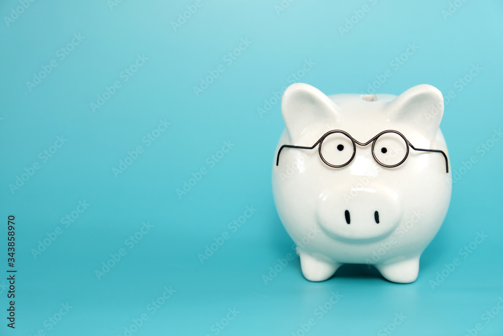 White ceramic piggy bank wearing reading glasses on blue teal background. Concept for money savings plan for retirement, aged society, or financial accounting