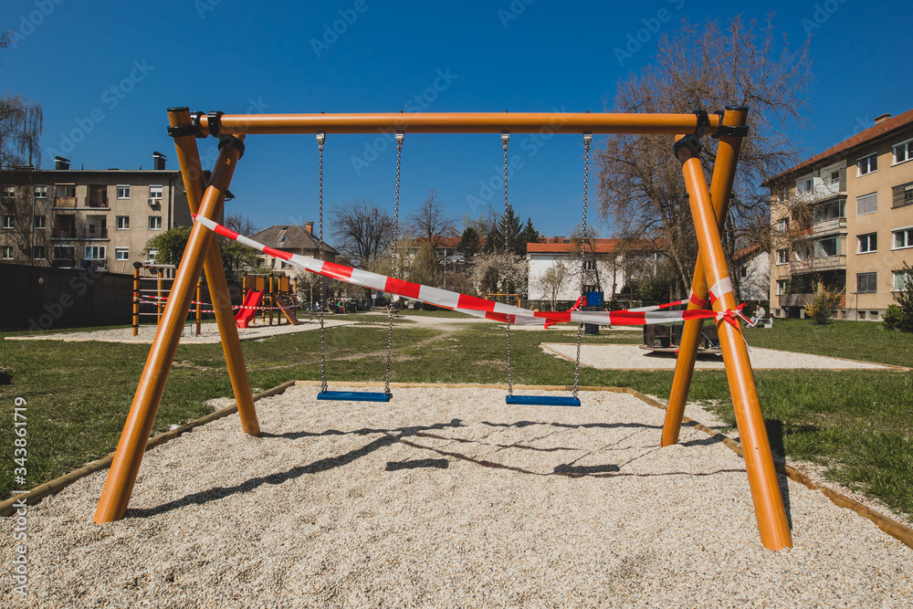 Closed children playground or swing on a chain due to corona virus restrictions taking in effect. Red and white tape over children swing. Closed children playground or swing on a chain due to corona virus restrictions taking in effect. Red and white tape over children swing.