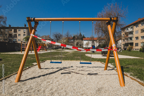 Closed children playground or swing on a chain due to corona virus restrictions taking in effect. Red and white tape over children swing.