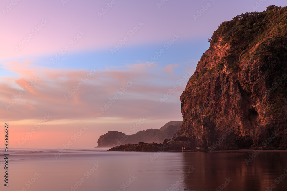 Piha, New Zealand, at sunset. Lion Rock (foreground) towers over the black sand beach