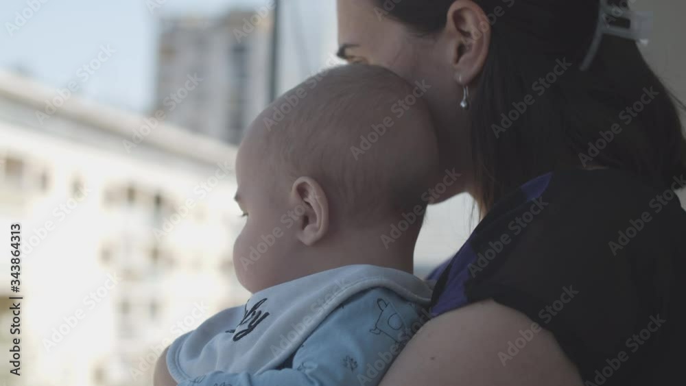 Mother holding a son in hands while standing on balcony and looking around, city buildings in the blurred background, sunny day. Mother holding a son in hands while standing on balcony and looking around, city buildings in the blurred background, sunny day.