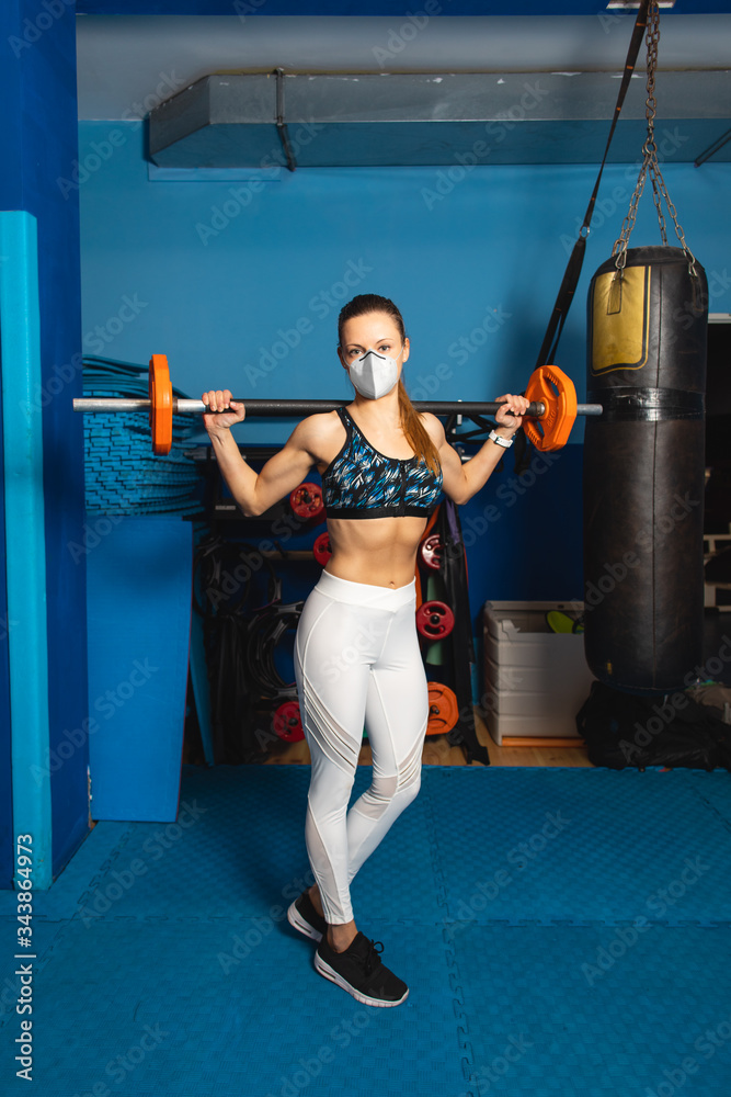 Young fit woman with n95 face mask  exercising at the gym with weights. Fitness strength workout under coronavirus health crisis.