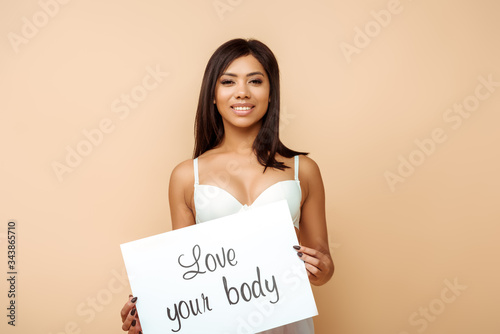 cheerful african american girl holding placard with love your body lettering isolated on beige