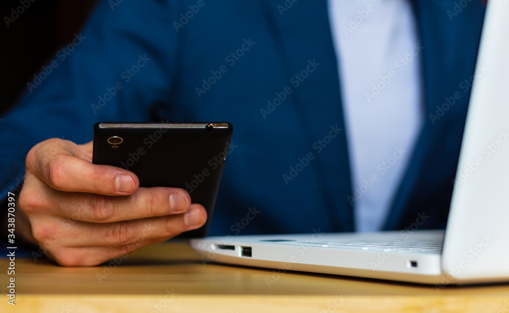 Close-up of the hands of a well-dressed man in a blue suit and a white shirt looking at his smartphone, using a laptop on a desk. Close-up of the hands of a well-dressed man in a blue suit and a white shirt looking at his smartphone, using a laptop on a desk.