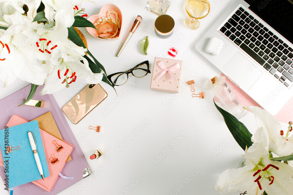 Creative colorful female workspace with laptop computer, glasses, cosmetics, flowers, colorful notebooks, golden clips on white desk. Flat lay, top view