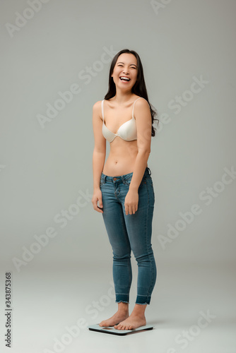 happy asian woman standing on scales and laughing on grey