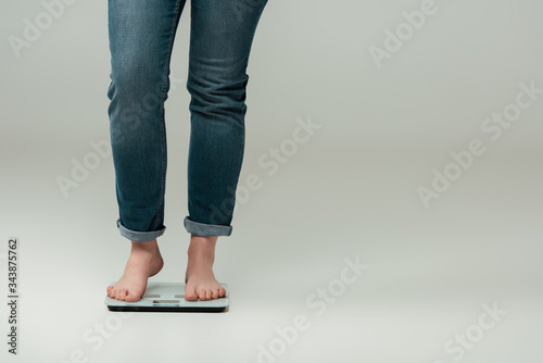cropped view of plus size girl in jeans standing on scales on grey