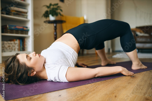 Beautiful plus size young woman practicing yoga, doing bridge pose, lying on mat with feet on floor, bending knees and pushing tailbone upward, stimulating abdominal organs, stretching spine and chest