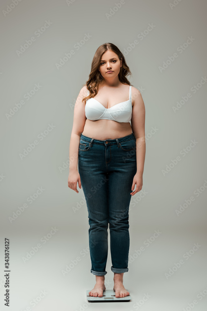 plus size girl in jeans and bra standing on scales and looking at camera on grey plus size girl in jeans and bra standing on scales and looking at camera on grey