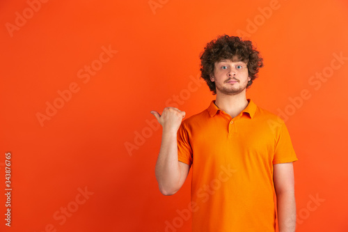 Pointing at side. Caucasian young man s monochrome portrait isolated on orange studio background. Beautiful male curly model in casual style. Concept of human emotions  facial expression  sales  ad.