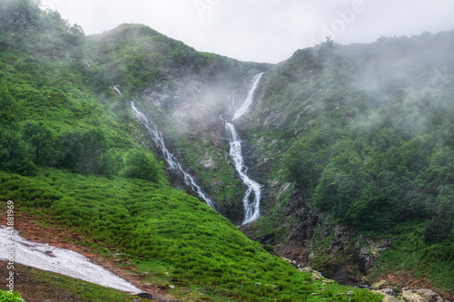 Green mountain range with a waterfall and not melted snow in spring or summer. A mountain range with a waterfall is covered in fog and clouds.