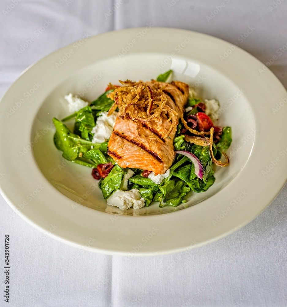 Stacked grilled salmon with onion rings salad, vegetable, and sauce on white plate Stacked grilled salmon with onion rings salad, vegetable, and sauce on white plate