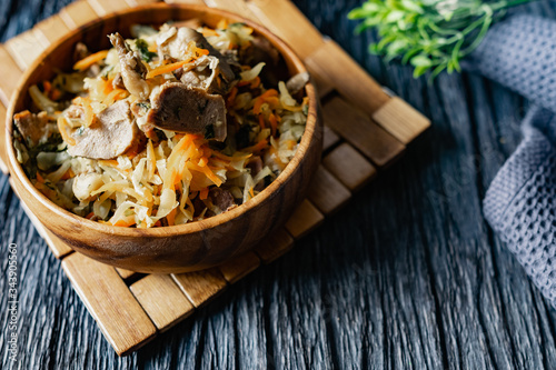 Stewed cabbage with oyster mushrooms and pork tongue with carrots and onions. Hot dish in a wooden plate on a wooden stand, next to a fork, a sprig of grass and a napkin. Black background with relief