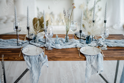 Romantic table setting for holiday dinner, wooden table served with dried flower, plates, golden cutlery, white candeles, bright dusty blue runner. Selective focus. photo