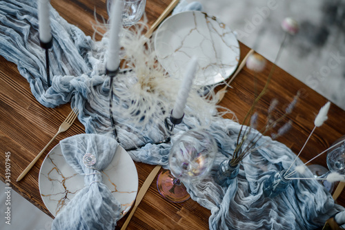 Minimalistic table setting for holiday dinner, wooden table with dried flower, plates, golden cutlery, white candeles, bright blue table runner. Selective focus. Top view