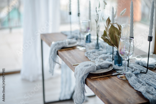 Minimalistic table setting for holiday dinner, wooden table with dried flower, plates, golden cutlery, white candeles, bright blue table runner. Selective focus.