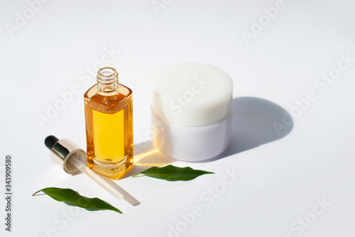 Yellow glass with a dropper. Bottle mockup cosmetic oil on white background with green leaves white creme. Copy space