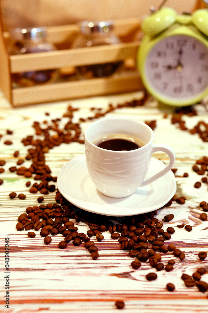 A cup of coffee on a platter with lots of sprinkled coffee beans. Alarm clock arrows at 9 am. Wooden textured table. Shy pastel background. Time to wake up in the morning. Natural food concept A cup of coffee on a platter with lots of sprinkled coffee beans. Alarm clock arrows at 9 am. Wooden textured table. Shy pastel background. Time to wake up in the morning. Natural food concept