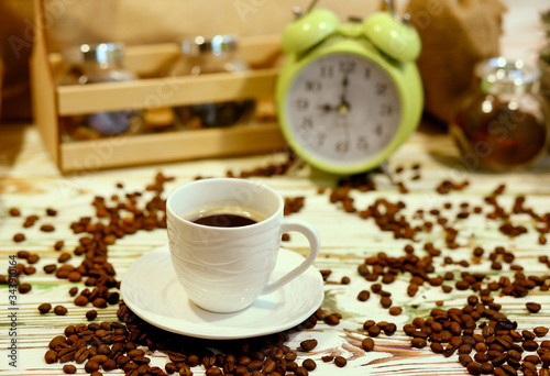 A cup of coffee on a platter with lots of sprinkled coffee beans. Alarm clock arrows at 9 am. Wooden textured table. Shy pastel background. Time to wake up in the morning. Natural food concept