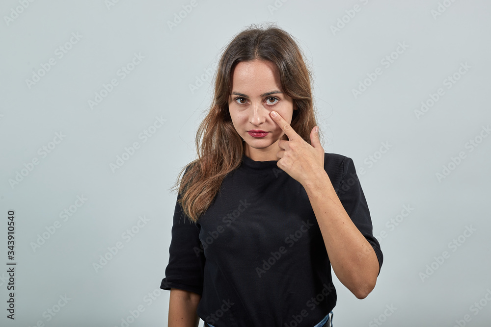 Cute young brunette woman in black t-shirt, blue jeans with belt on gray background, beautiful girl shows her finger on the eye that she has vision problems Cute young brunette woman in black t-shirt, blue jeans with belt on gray background, beautiful girl shows her finger on the eye that she has vision problems