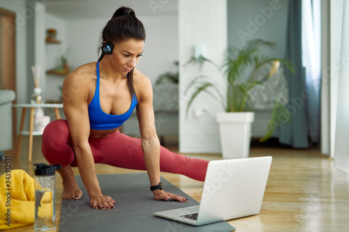 Young muscular build woman doing stretching exercises while using laptop at home. © Drazen