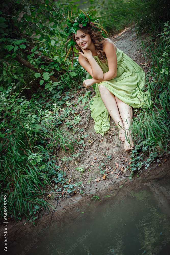 Beautiful young woman in a green dress and a wreath in a sunny forest
